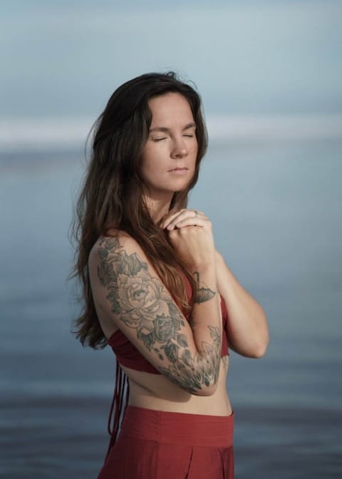 Frau am Strand, seitlich fotografiert. Beide Hände auf der Brust. Offene lange Haare. Augen geschlossen. Rote lange Hose und bauchfreies Top.