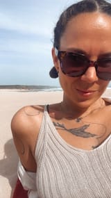 Eine Frau am Strand, sitzend. Beide arme hinter ihr aufgestützt. Hat eine Sonnenbrille auf und die Haare zu zwei Braids geflochten am Kopf. Sie lächelt fröhlich.