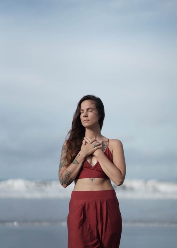 Eine Frau am Strand, sie trägt eine rote Hose und ein rotes Bauchfreies Oberteil, hat die Hände auf ihr Herz gelegt, Augen geschlossen, der Wind fegt durch die Haare.